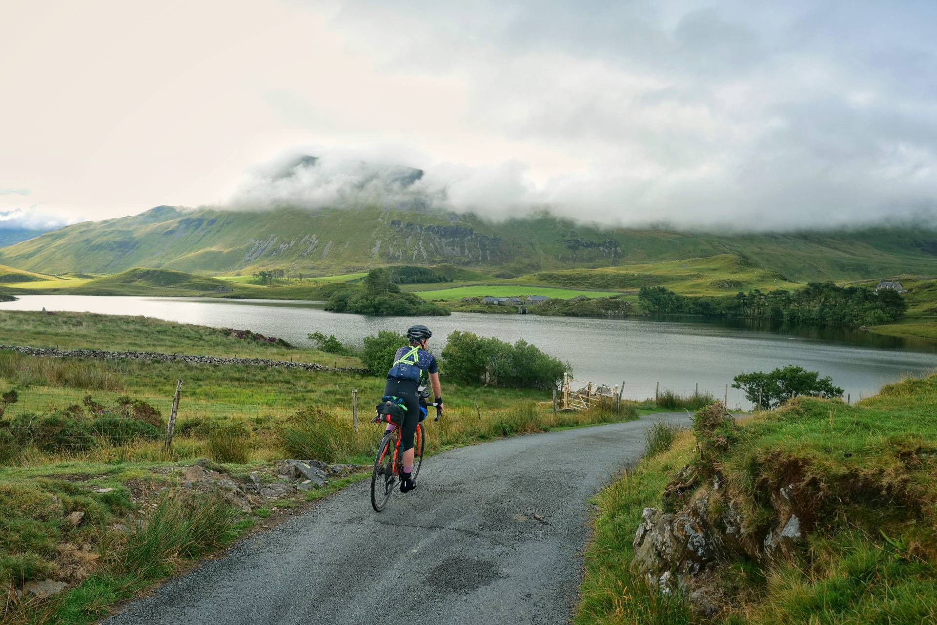 Rider on a lakeside road beneath cloud-capped mountains during The Perfidious Albion